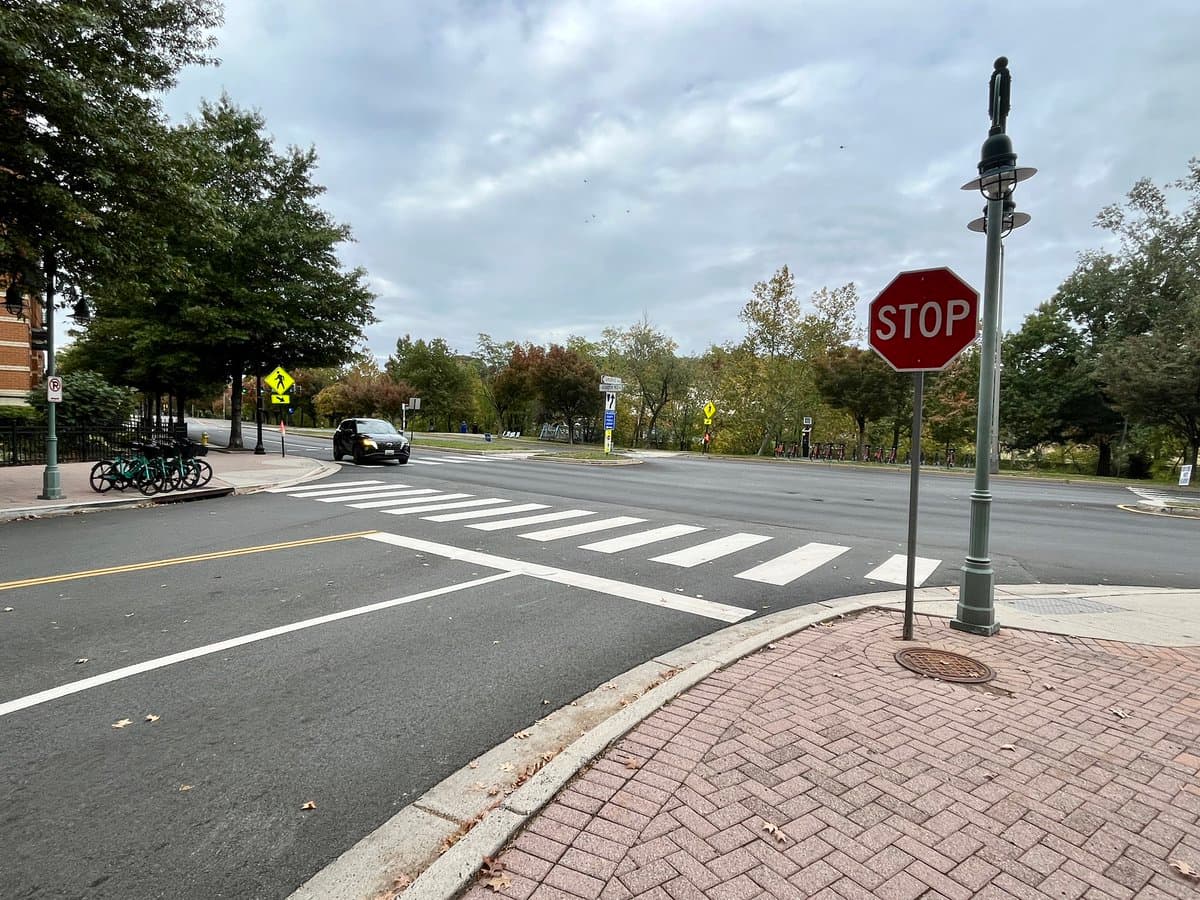 A street-level view of a city intersection on an overcast day, featuring a stop sign, a crosswalk, and a row of parked rental e-bikes on the left sidewalk.