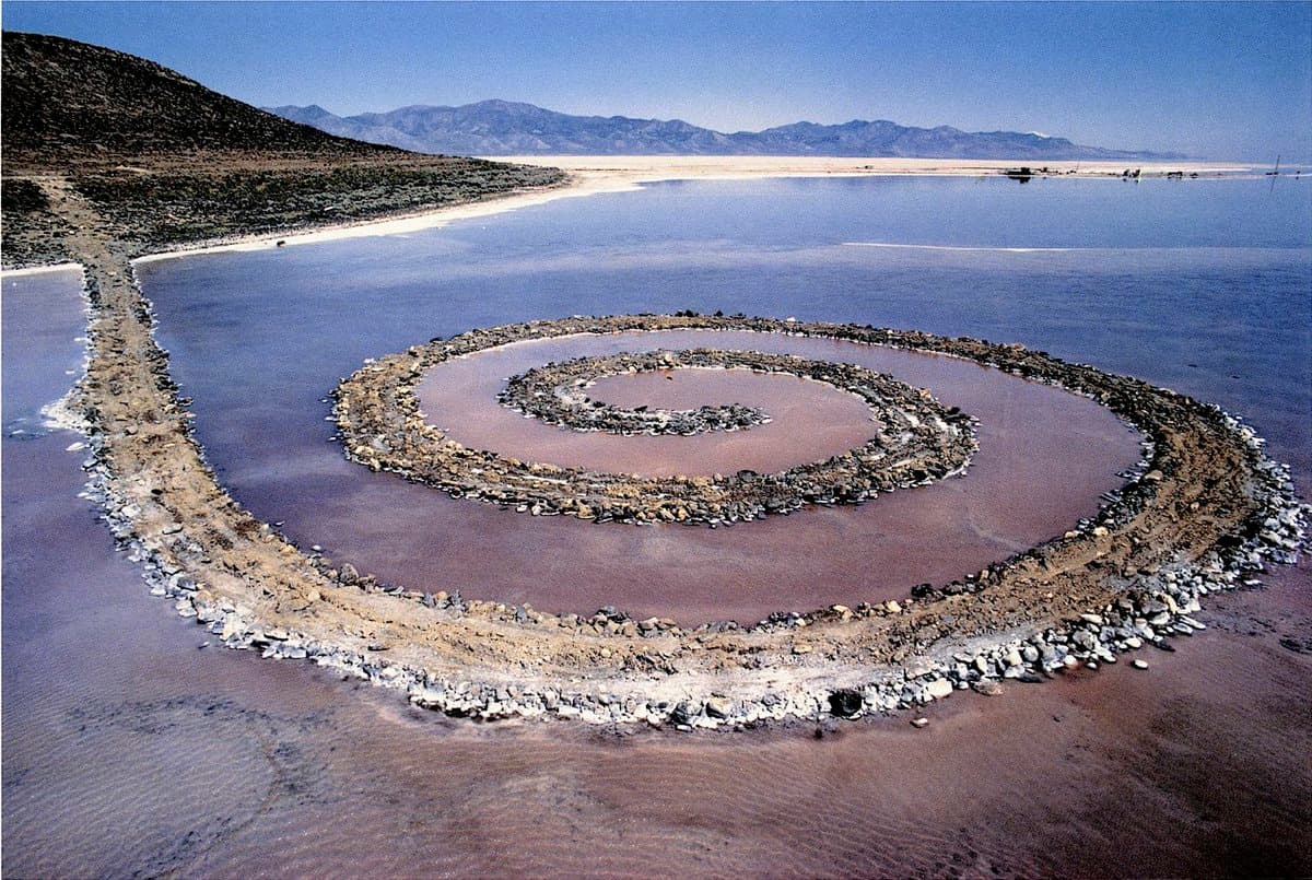 An aerial photograph of the Spiral Jetty, a large land art installation consisting of a spiral-shaped jetty made of rocks and earth extending into a shallow body of water. The structure is set against a backdrop of a dry, hilly landscape and distant mountains under a clear blue sky.