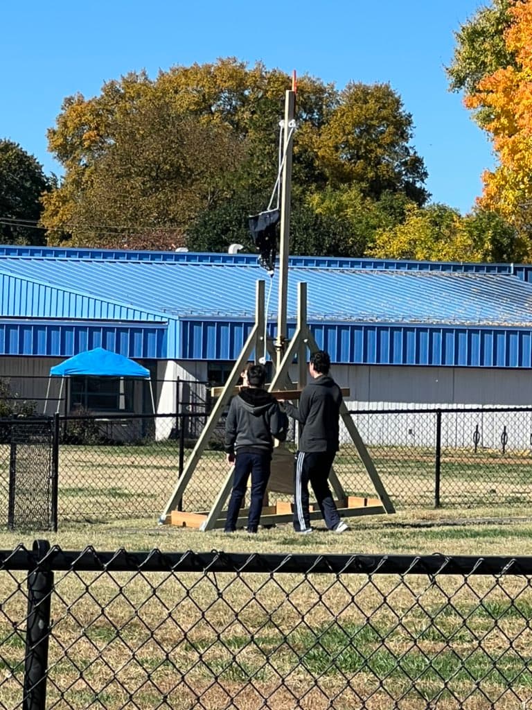 Two people stand in a grassy field operating a large wooden trebuchet, with a blue-roofed building and autumn trees in the background.