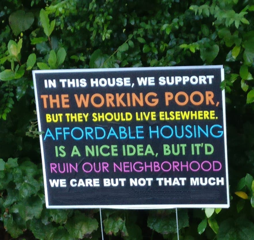 A black yard sign with colorful text is displayed in front of a green hedge. The sign contains a satirical statement about housing and community attitudes.