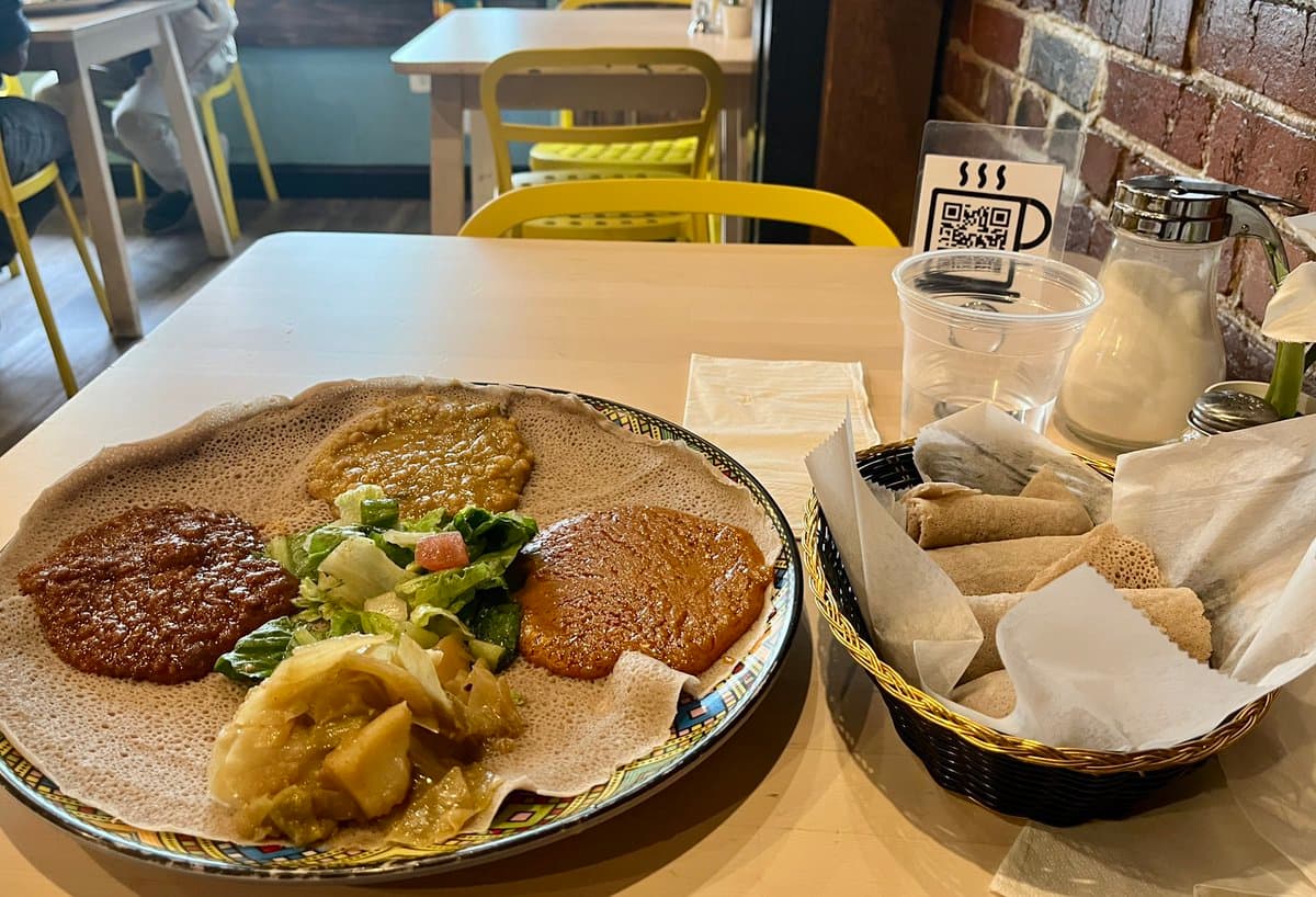 A plate of Ethiopian food featuring injera topped with various stews and a side salad, served next to a basket of rolled injera wraps in a restaurant setting.