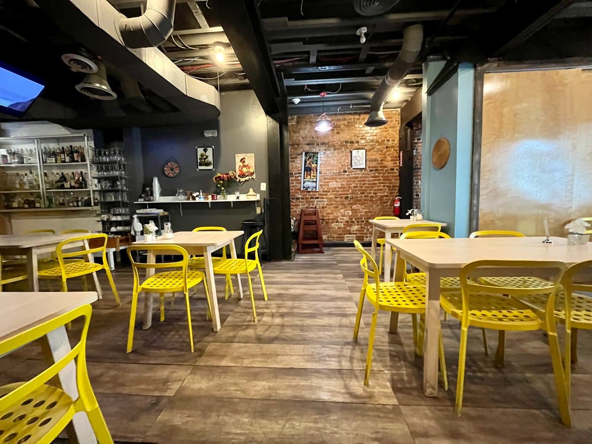A wide interior shot of a restaurant dining area featuring white tables and bright yellow chairs on a tiled floor. The space has an industrial aesthetic with exposed brick walls, a bar area with shelves of bottles, and an open ceiling with visible ductwork.