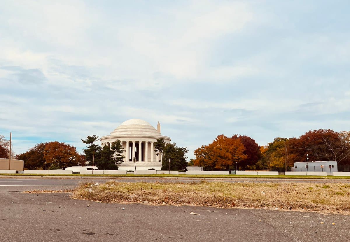 A wide shot of the Jefferson Memorial in Washington D.C. on a cloudy day, surrounded by trees with autumn foliage and a paved road in the foreground.