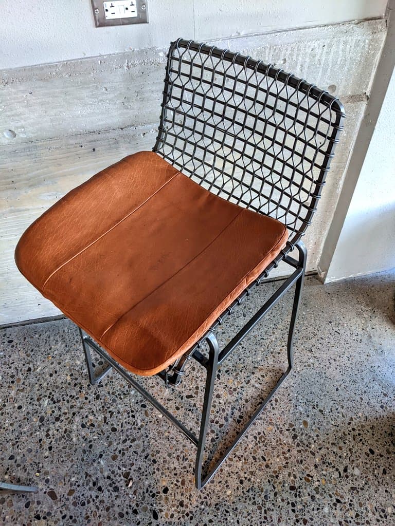 A photograph of a chair with a black wire mesh back and a brown leather seat cushion, positioned on a terrazzo floor against a concrete wall. The chair is the central subject, shown from a slightly elevated angle.
