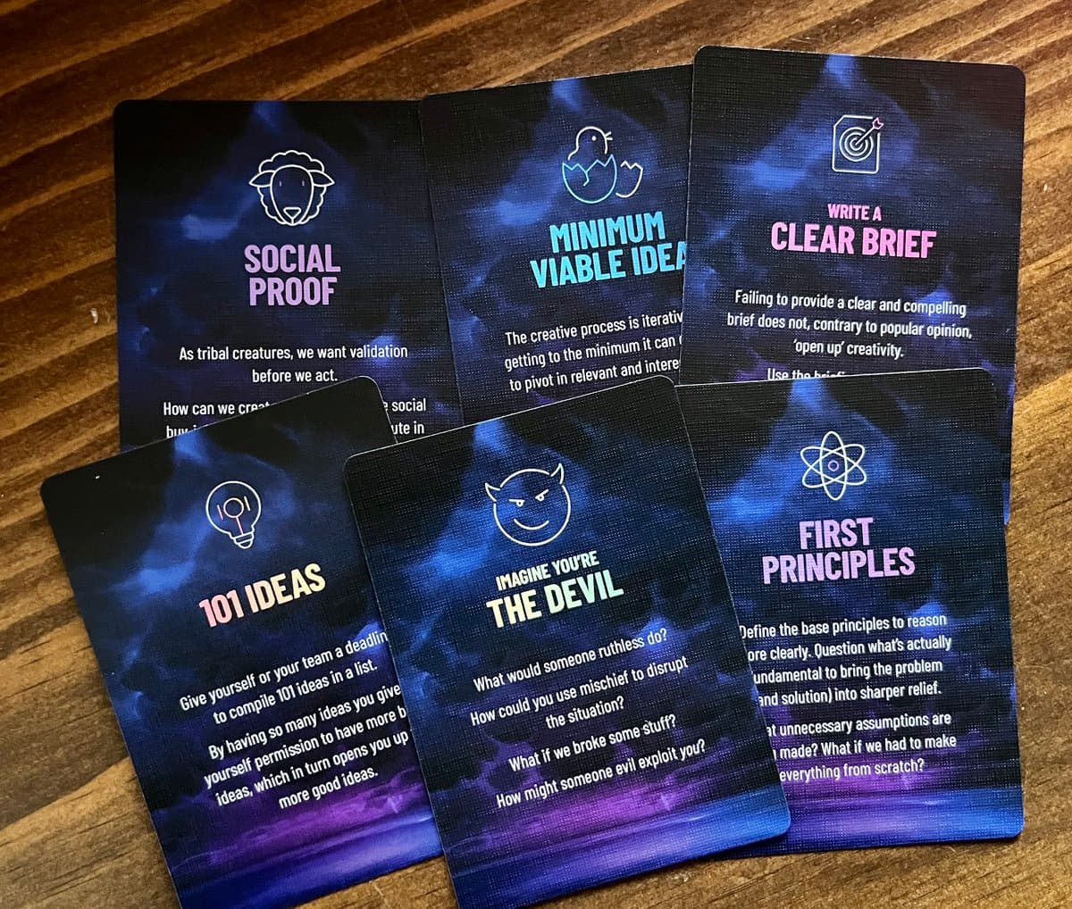 A photograph of six printed brainstorming cards with a dark blue and purple cosmic background, arranged on a wooden surface. The cards feature white line-art icons and text prompts for creative problem-solving, including 'Social Proof,' 'Minimum Viable Idea,' 'Write a Clear Brief,' '101 Ideas,' 'Imagine You're the Devil,' and 'First Principles'.