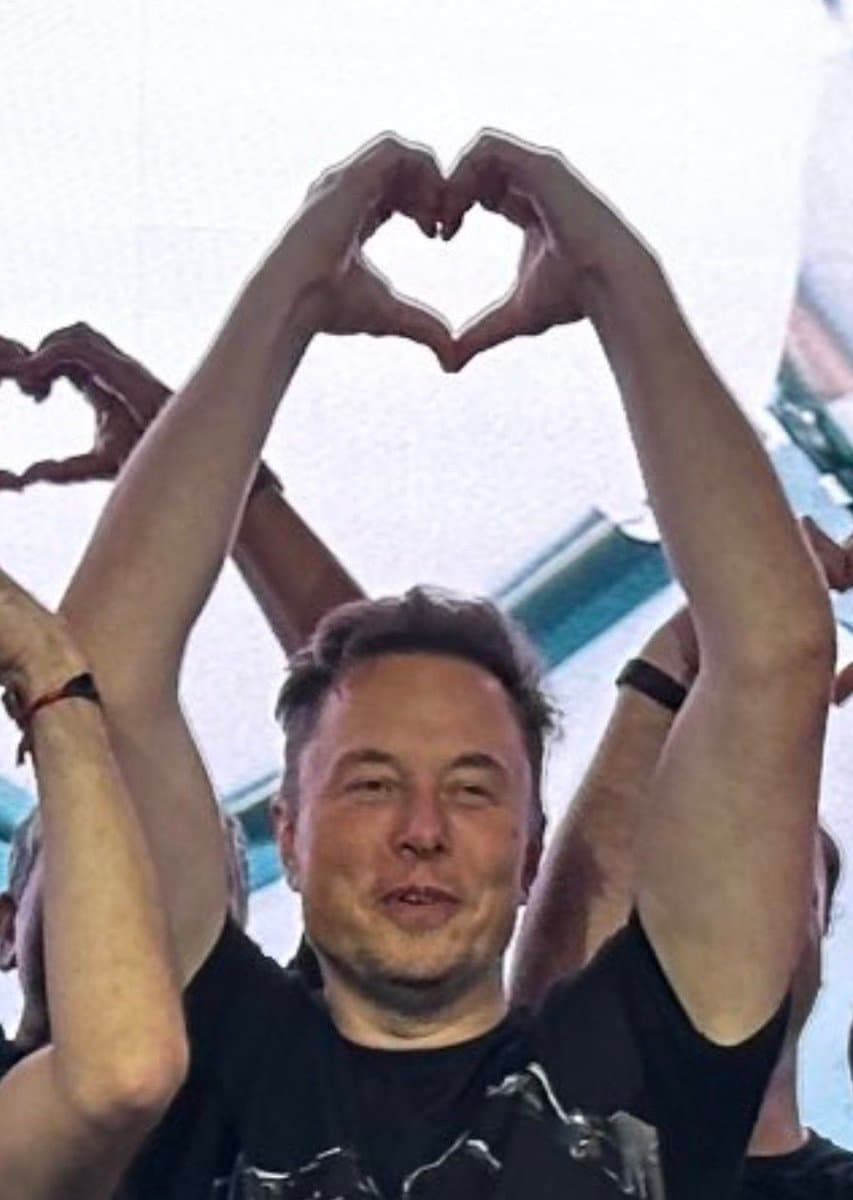 A photograph of a man in a black t-shirt raising both arms to form a heart shape with his hands, with other people in the background making similar gestures.
