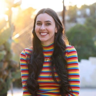 A portrait of a smiling woman with long, dark wavy hair wearing a brightly colored, horizontally striped turtleneck sweater. She is standing outdoors with a blurred background of trees and foliage illuminated by warm, golden sunlight.
