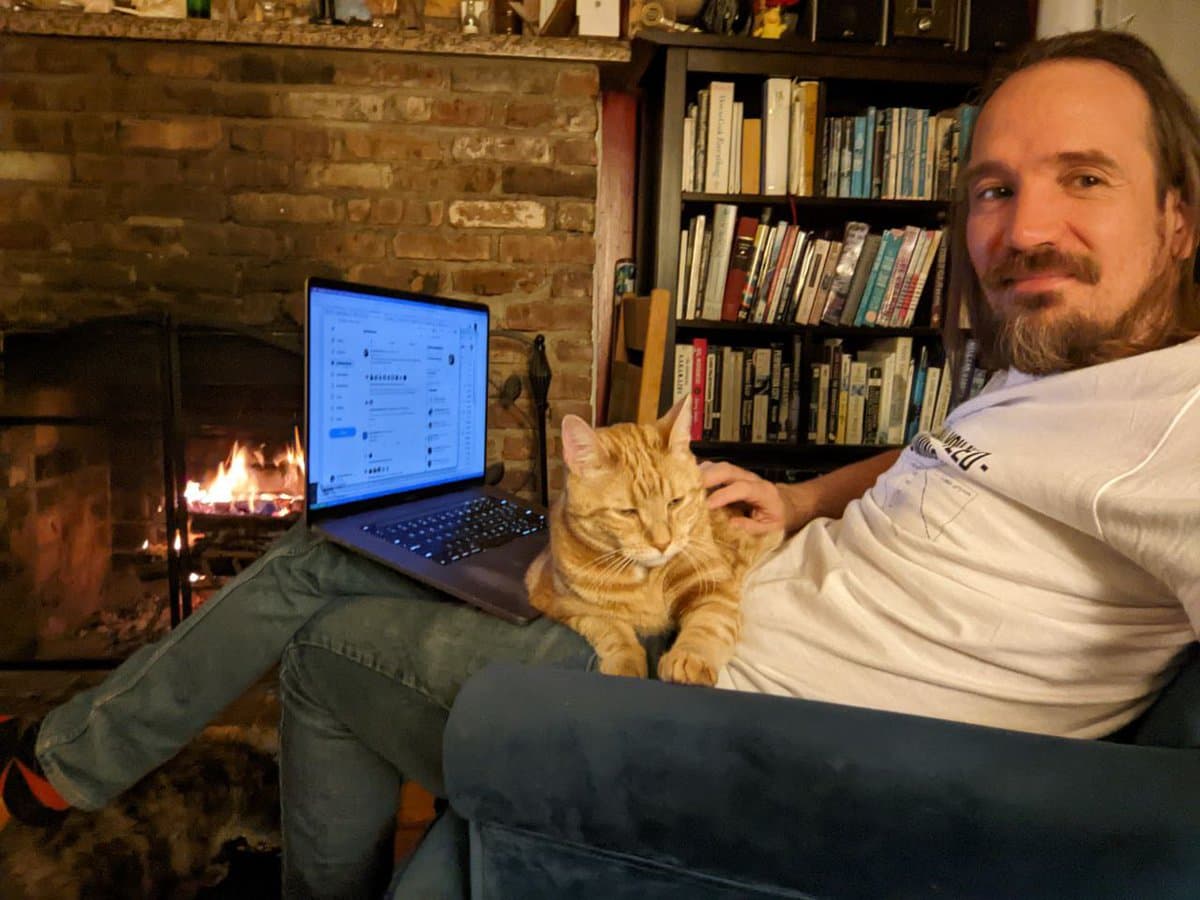 A man with a beard sits in a blue armchair next to a lit brick fireplace, holding an orange tabby cat on his lap while a laptop rests on his legs. A bookshelf filled with books is visible in the background.