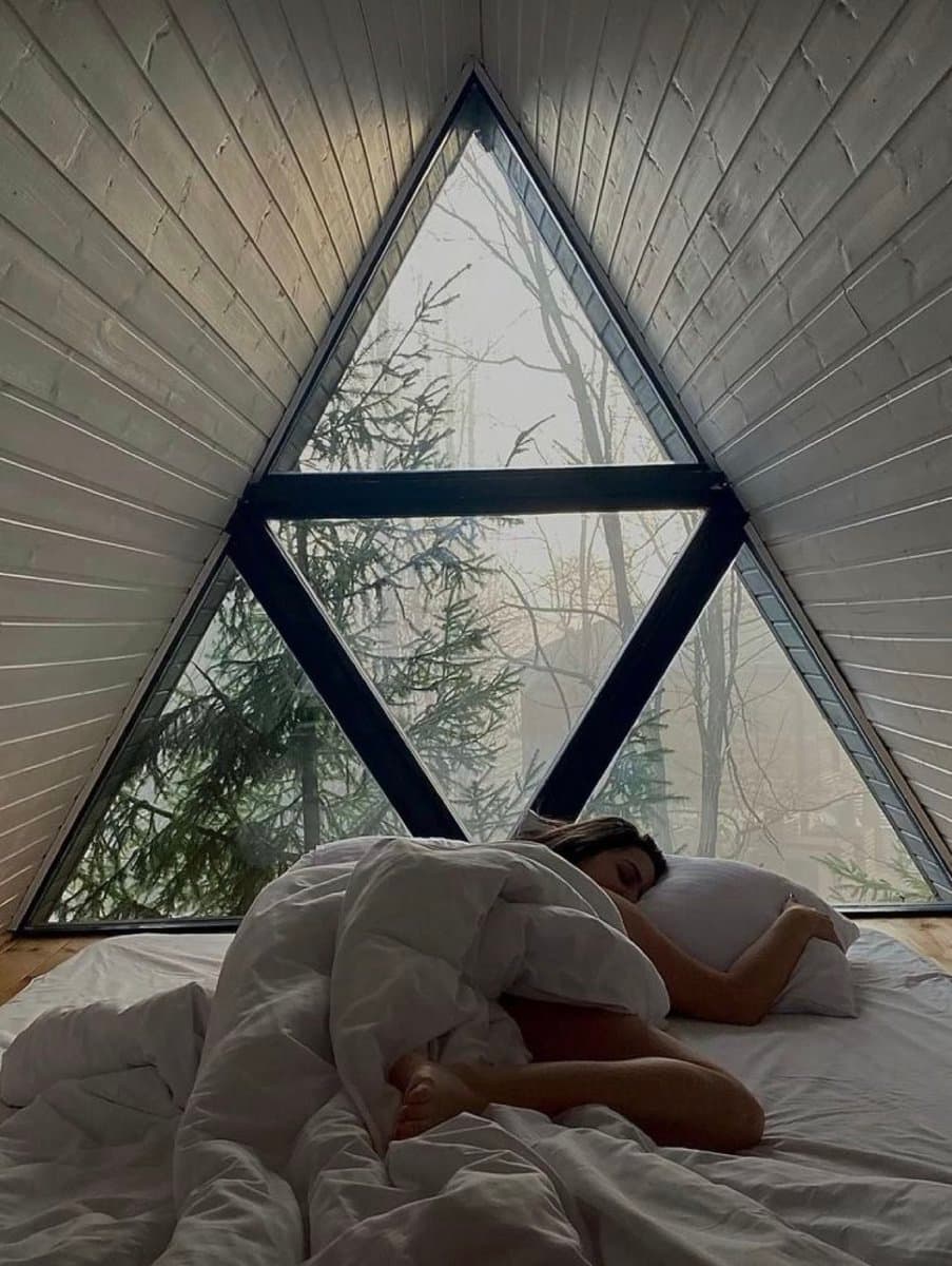 A person is sleeping in a white bed inside a small A-frame loft, with a large triangular window behind them looking out onto a misty forest.