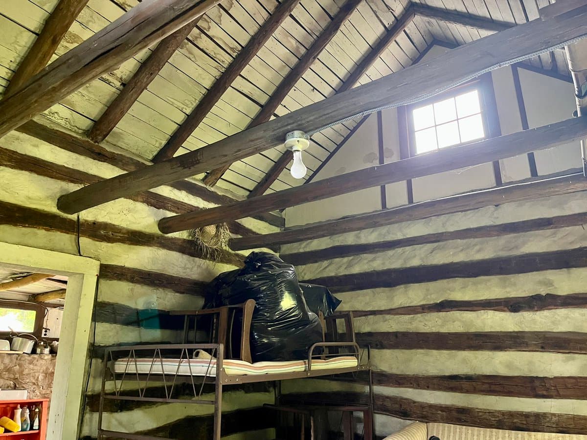 A photo of a rustic attic room featuring exposed wooden beams, a sloped ceiling, and a small window. A metal bunk bed with a mattress is positioned against a wall with horizontal wooden planks, and a large black trash bag sits on the upper bunk.
