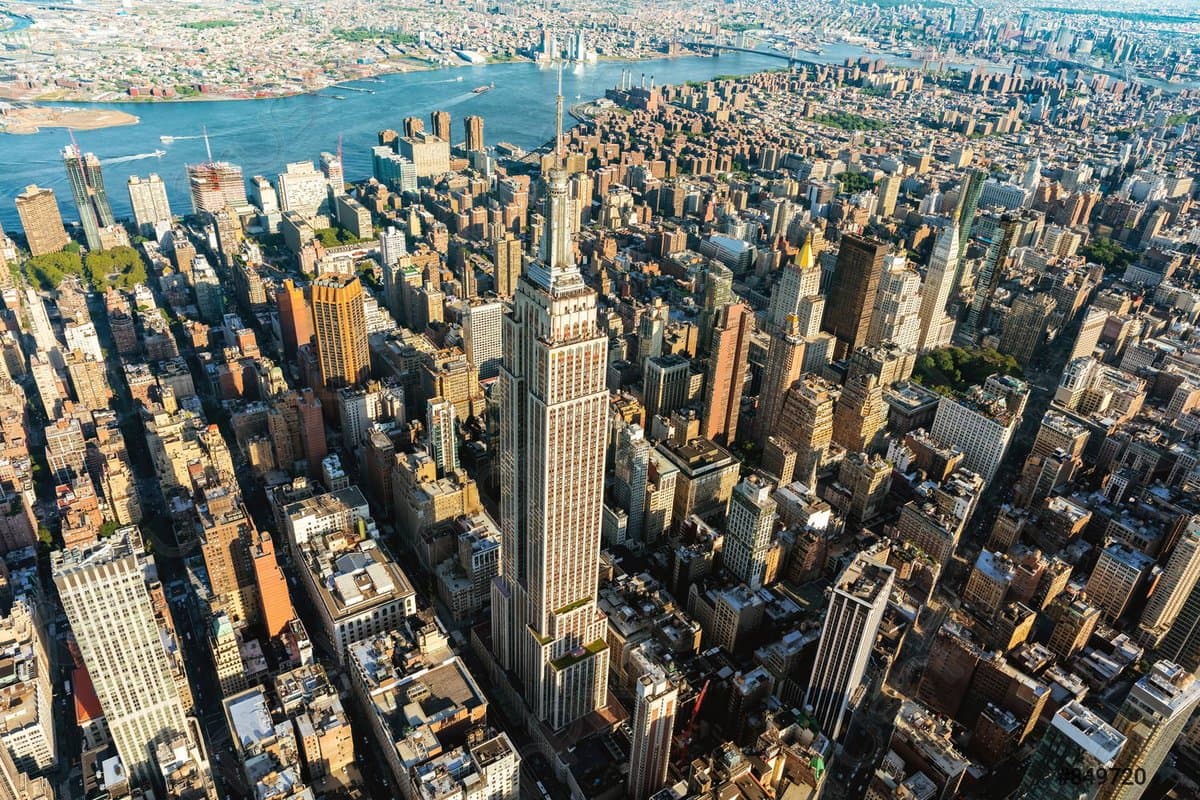 An aerial photograph of a dense city skyline featuring the Empire State Building prominently in the center, surrounded by numerous other skyscrapers and a large river in the background. The image is a standard landscape photo with no visible text, charts, or document elements.
