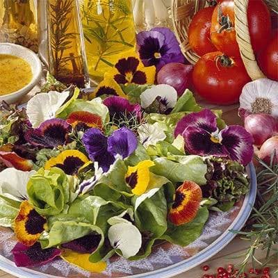 A close-up photograph of a fresh salad on a white plate, topped with colorful edible pansy flowers in shades of purple, yellow, and white. The background features ingredients including red tomatoes, red onions, a bottle of olive oil, and a small bowl of yellow dressing.