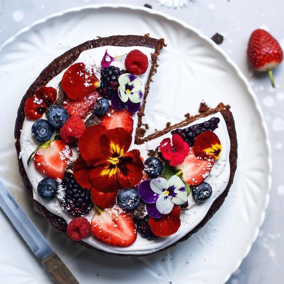 A top-down view of a decorated chocolate cake on a white plate, topped with white frosting, fresh berries (strawberries, blueberries, raspberries, blackberries), and colorful edible flowers. A slice has been cut from the cake, and a knife rests beside the plate.