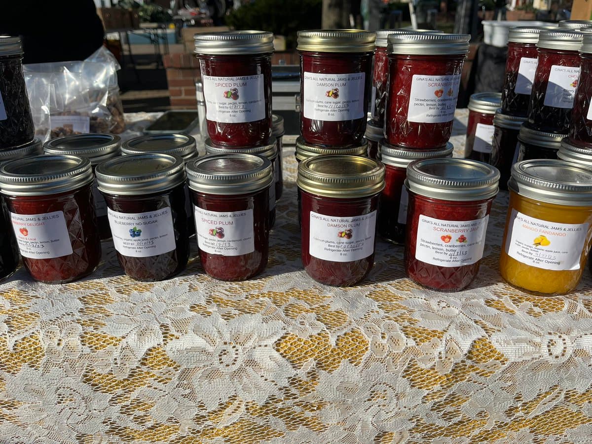 A display of numerous glass jars filled with various types of jam and jelly arranged on a table covered with a white lace tablecloth. The jars feature white labels with handwritten details, including flavors like Damson Plum, Spiced Plum, and Mango Fandango.
