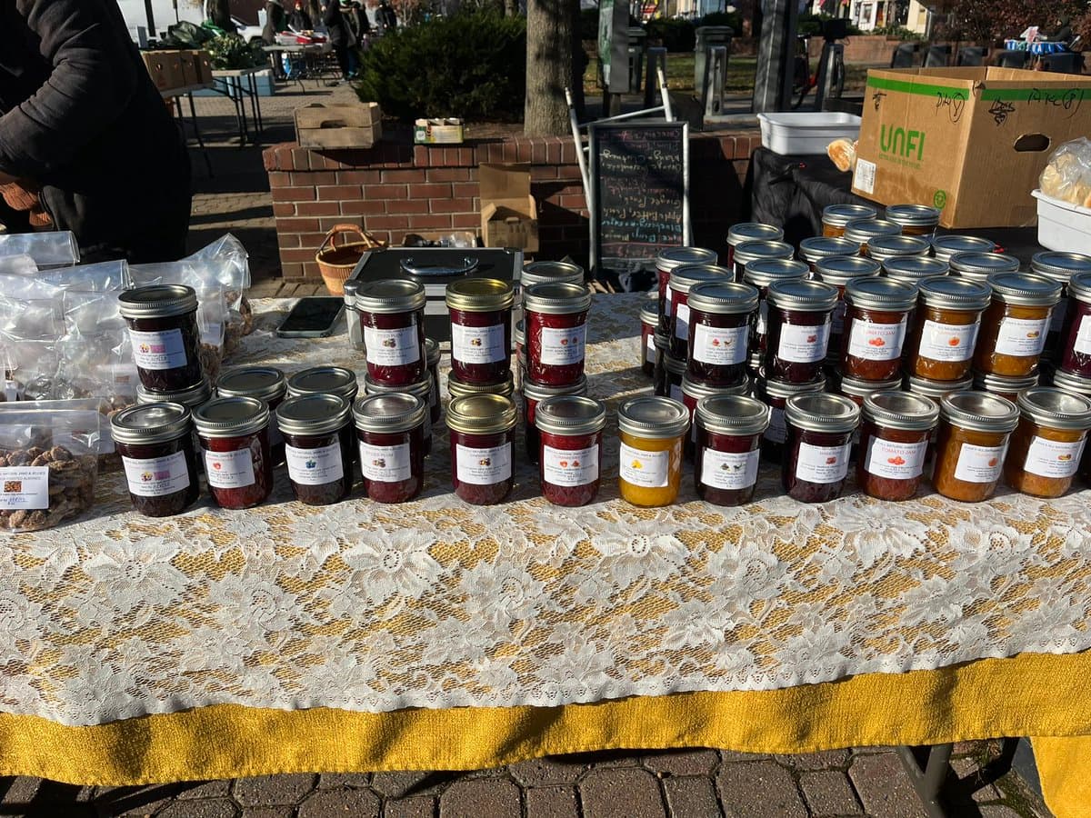 A market table covered in a white lace tablecloth displays numerous jars of jam in various colors, including red, purple, and orange. In the background, there is a cardboard box with a green 'UNFI' logo and a chalkboard sign.