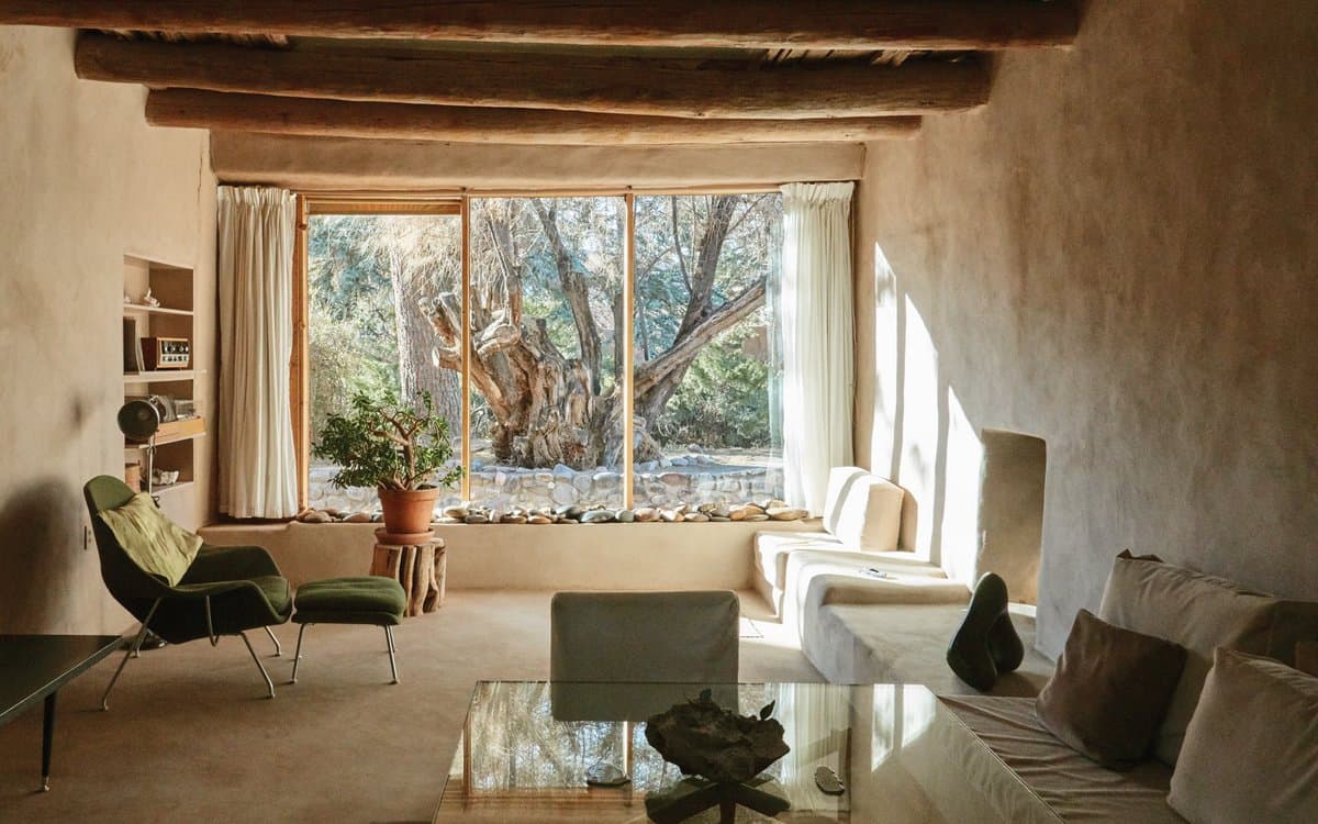 A photograph of a rustic, earth-toned living room featuring adobe walls, exposed wooden ceiling beams, and a large window overlooking a wooded landscape. The space is furnished with a green lounge chair, a white sofa, and a glass coffee table, creating a serene and natural atmosphere.