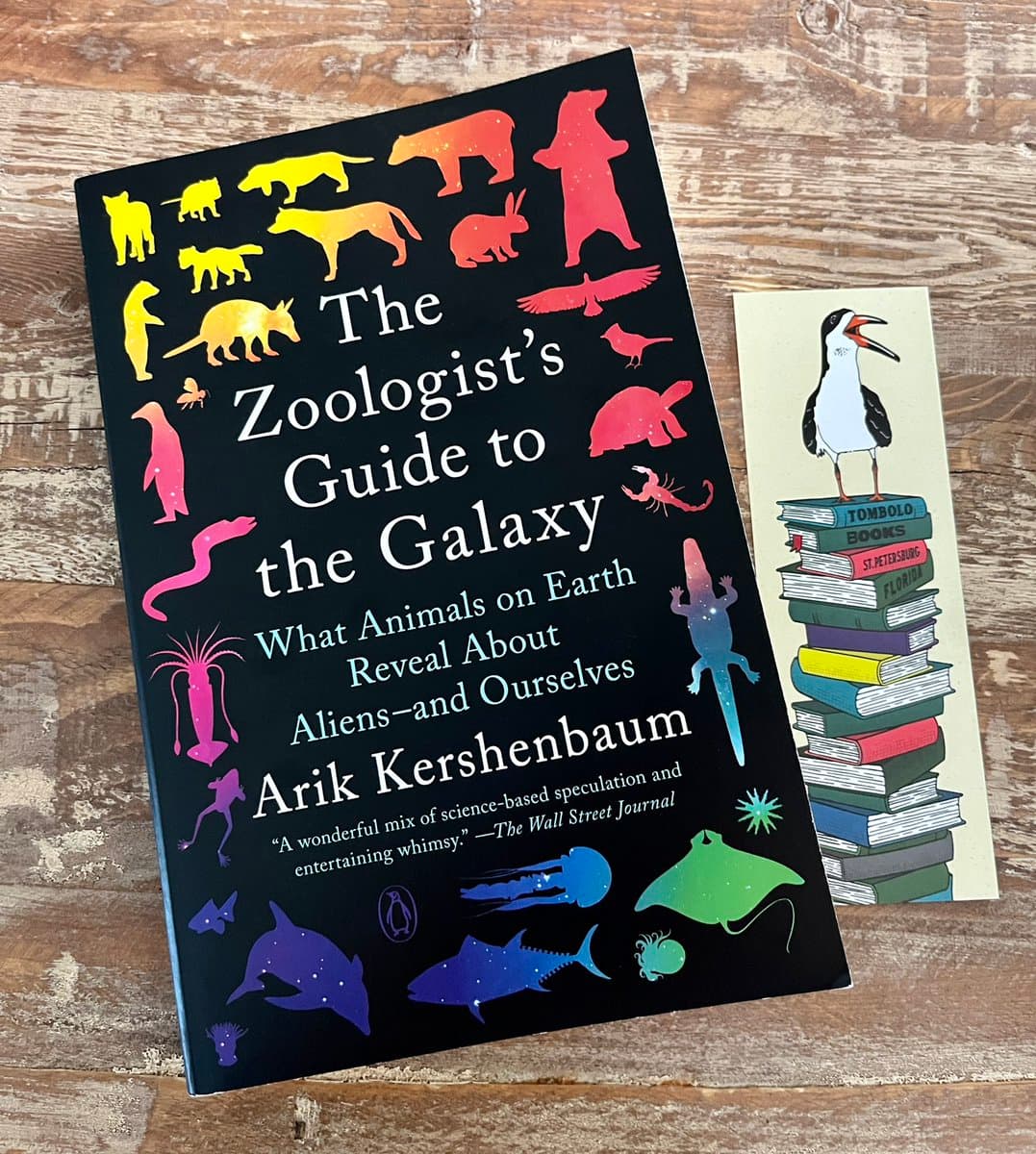 A photograph of the book 'The Zoologist's Guide to the Galaxy' by Arik Kershenbaum lying on a wooden surface next to a bookmark featuring a bird on a stack of books.