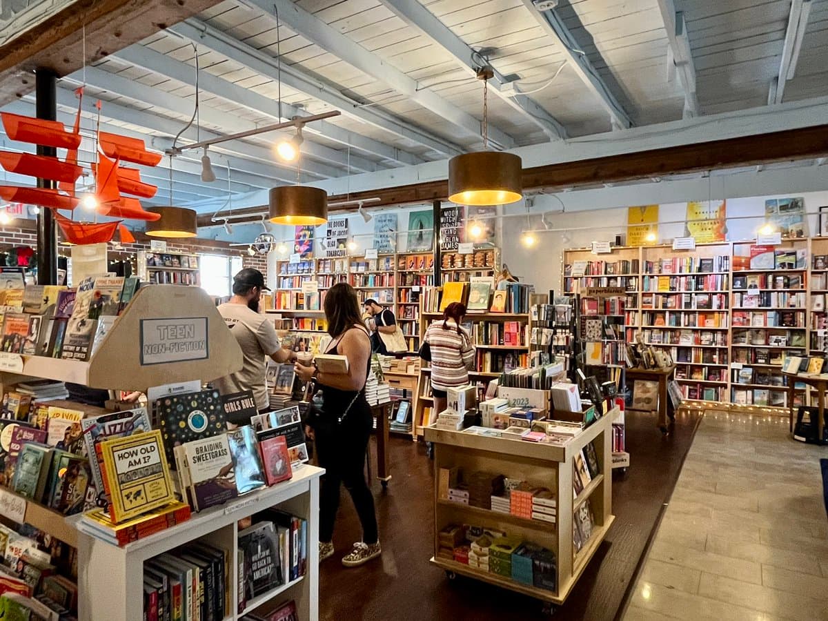 A wide-angle photograph of the interior of a bookstore, showing wooden bookshelves filled with books, display tables, and several customers browsing. A sign on a display stand in the foreground reads "TEEN NON-FICTION."