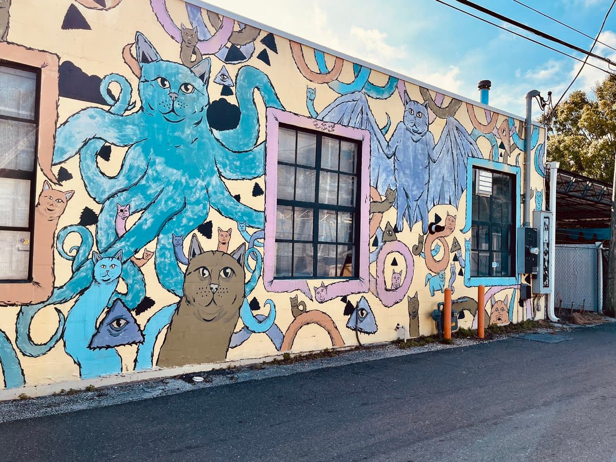 A photograph of a large, whimsical mural painted on the side of a building, featuring numerous cats in various forms, including a large blue octopus-cat and a winged cat. The scene is set on a street corner with a paved road in the foreground.
