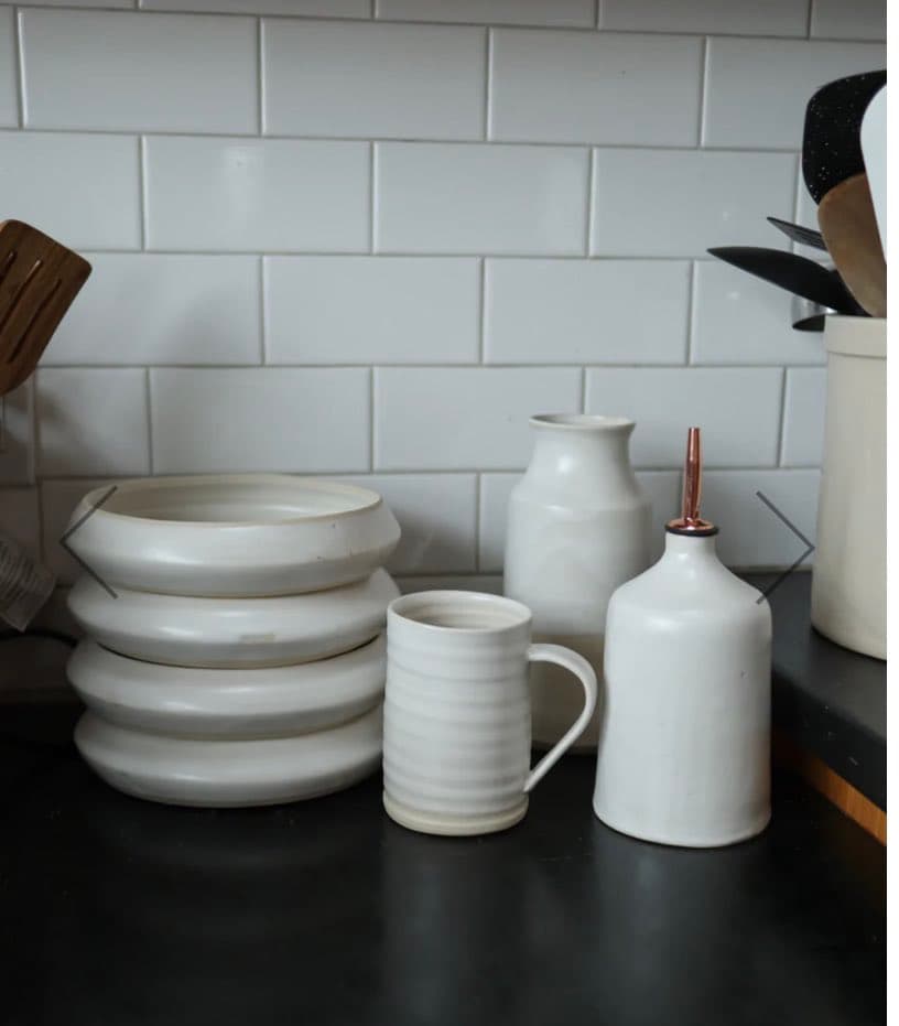 A collection of white ceramic kitchenware, including stacked plates, a mug, a pitcher, and a bottle with a copper pour spout, arranged on a dark countertop against a white subway tile backsplash.