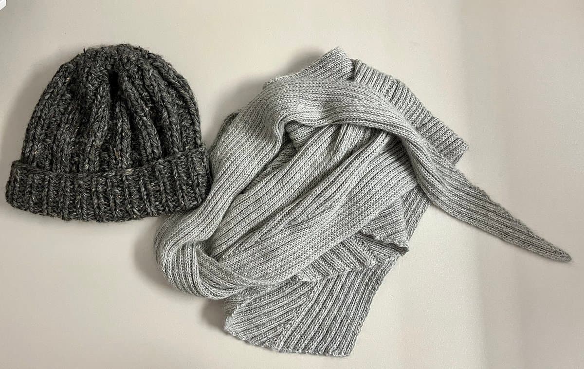 A flat-lay photograph of two hand-knitted items on a white surface: a dark gray ribbed beanie on the left and a light gray ribbed scarf draped beside it. The image showcases the texture and craftsmanship of the knitted pieces.