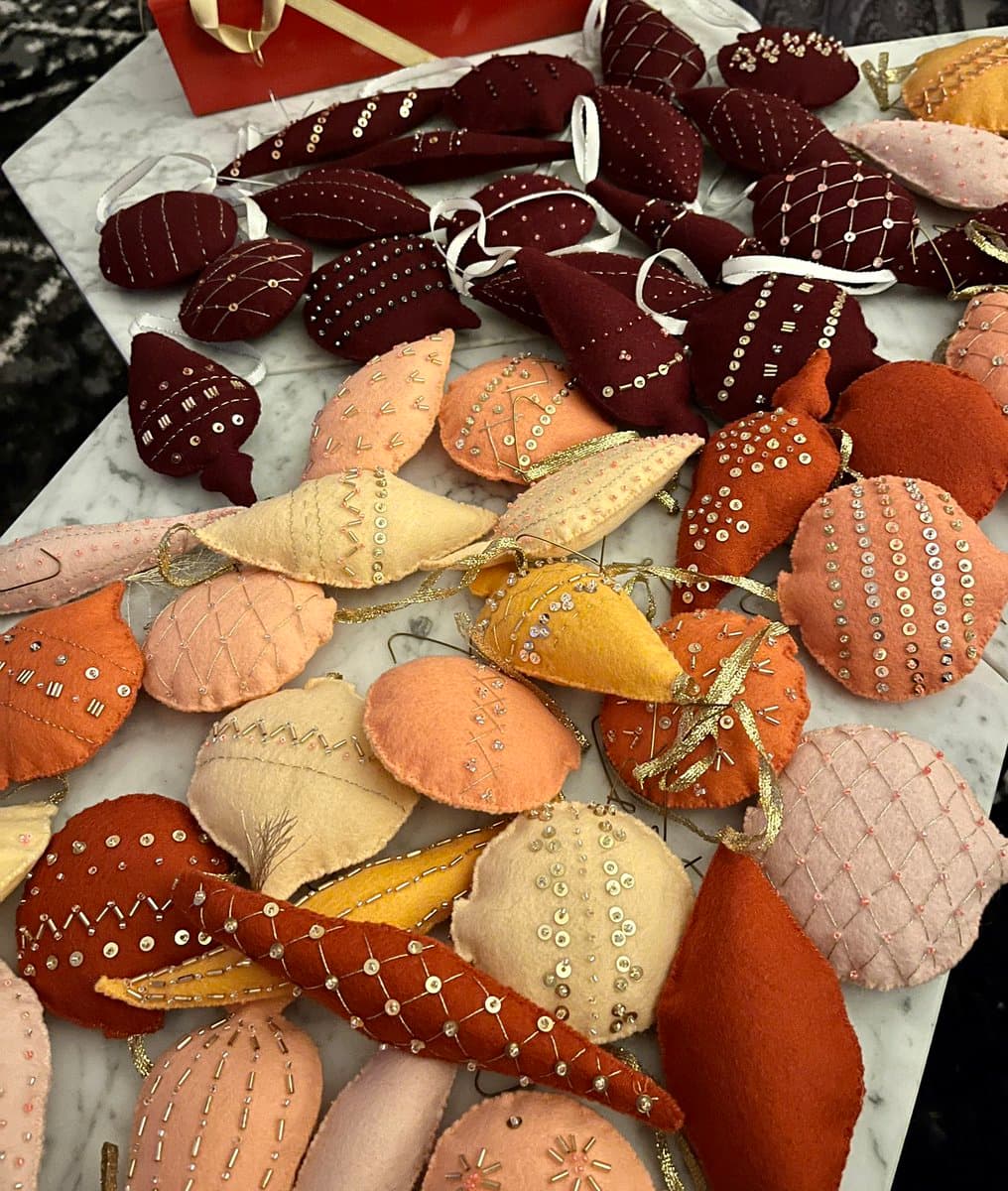 A collection of handmade felt ornaments in shades of burgundy, rust, and cream, decorated with decorative stitching and sequins, laid out on a white marble surface.