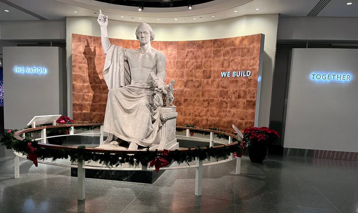 A large white marble statue of a seated man with one arm raised stands in the center of a modern, circular room. The statue is surrounded by a low railing decorated with a Christmas garland and red bows, with poinsettia plants placed nearby. Illuminated text on the surrounding walls reads "THE NATION," "WE BUILD," and "TOGETHER."