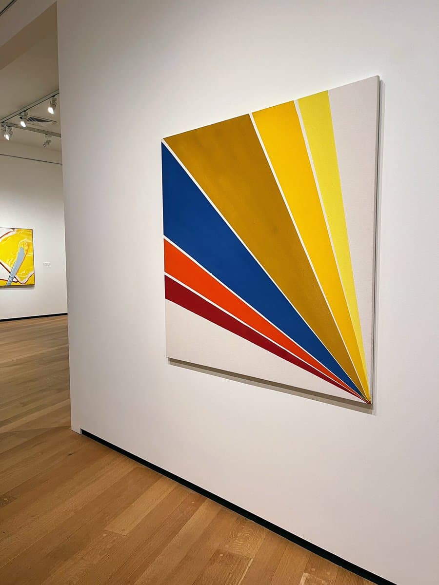 A photograph taken inside a gallery showing a large abstract painting with diagonal stripes of blue, yellow, and red on a white canvas, mounted on a white wall next to a wooden floor.