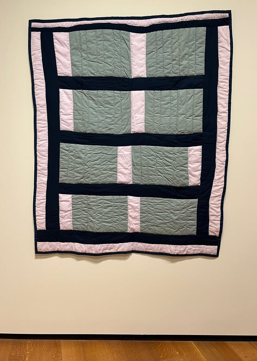 A photograph of a large, rectangular quilt hanging on a gallery wall. The quilt features a geometric pattern of sage green, pale pink, and dark navy blue fabric panels with visible quilting stitches.