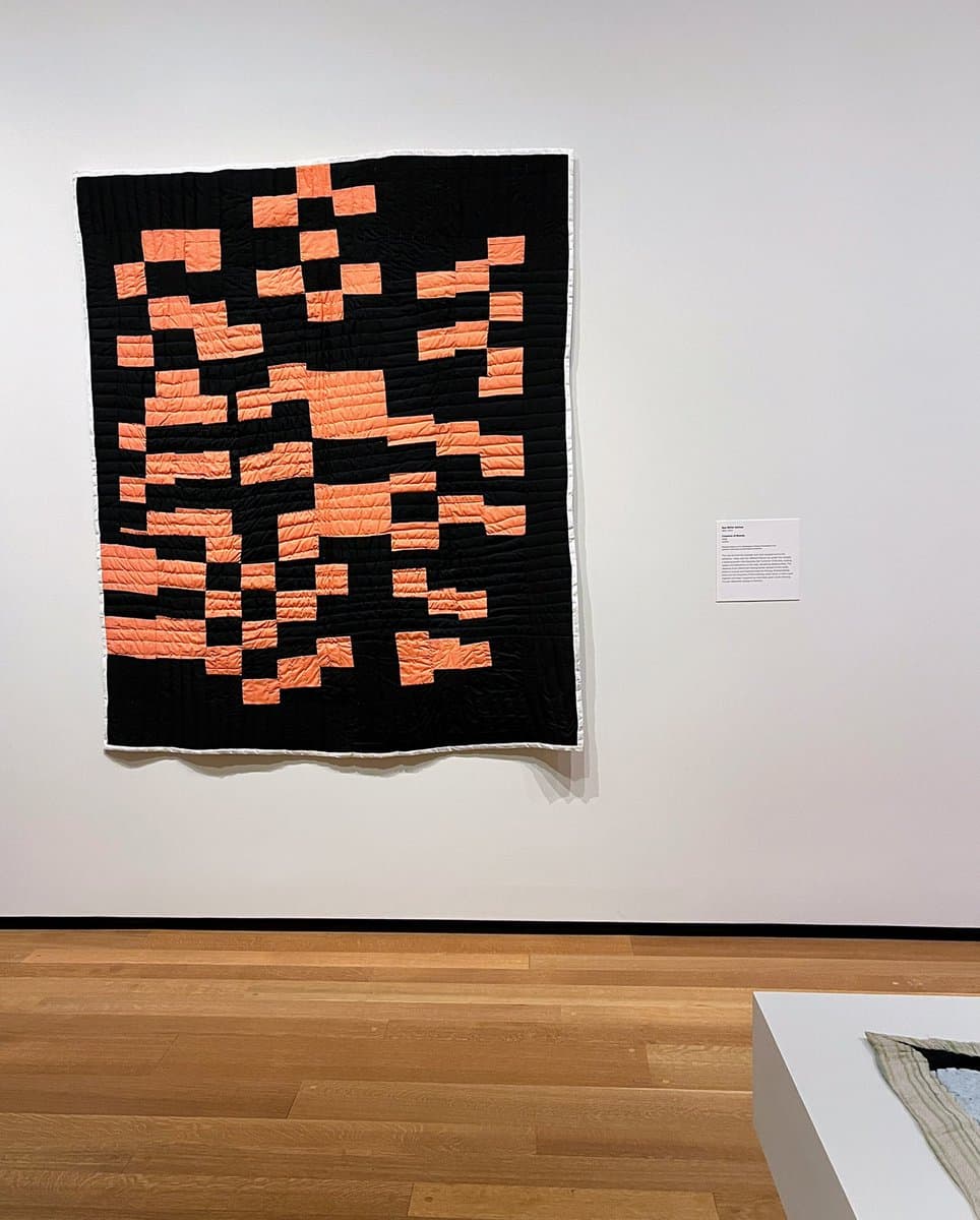 A large, black and orange pixelated quilt hangs on a white gallery wall, accompanied by a small informational placard to its right. The piece is displayed in a museum setting with a wooden floor and a white pedestal in the foreground.