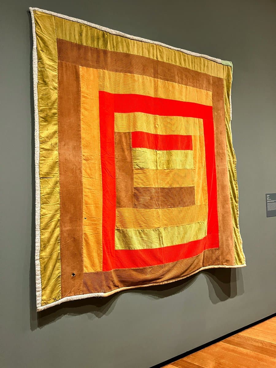 A large, square textile artwork featuring a geometric pattern of concentric rectangles in shades of red, yellow, and brown, displayed on a gray gallery wall.