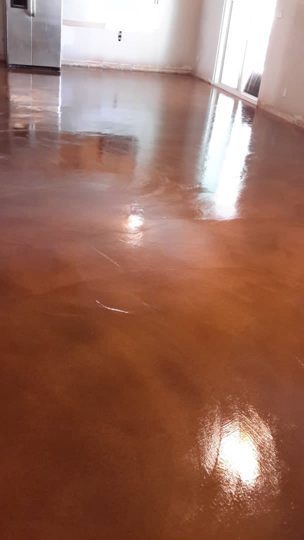 A photo of a room with a glossy, polished concrete floor that reflects the light from a window and a stainless steel refrigerator.