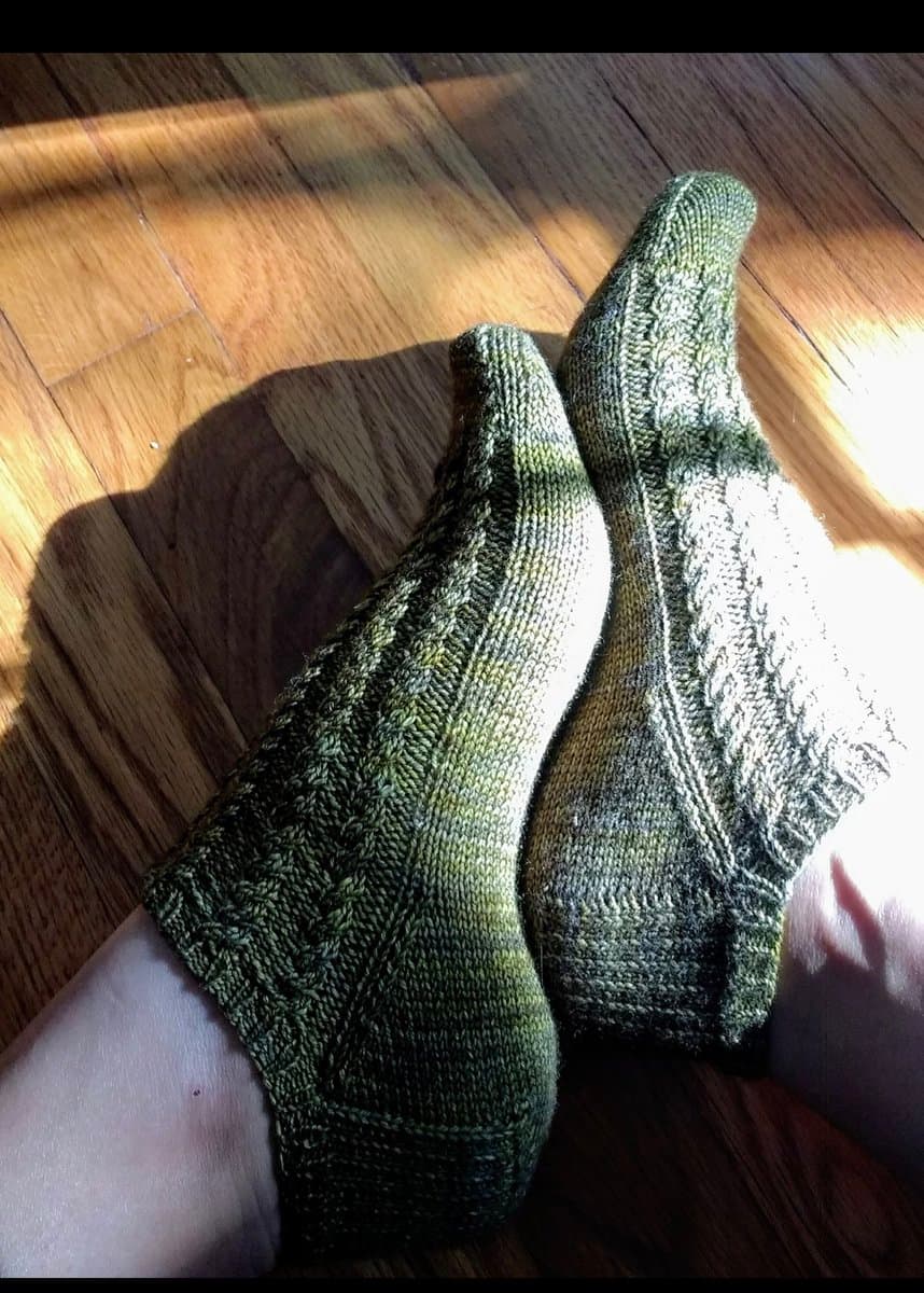 A pair of green, hand-knitted socks worn on a person's feet, resting on a wooden floor. The socks feature a textured, cable-knit pattern and are illuminated by bright sunlight.