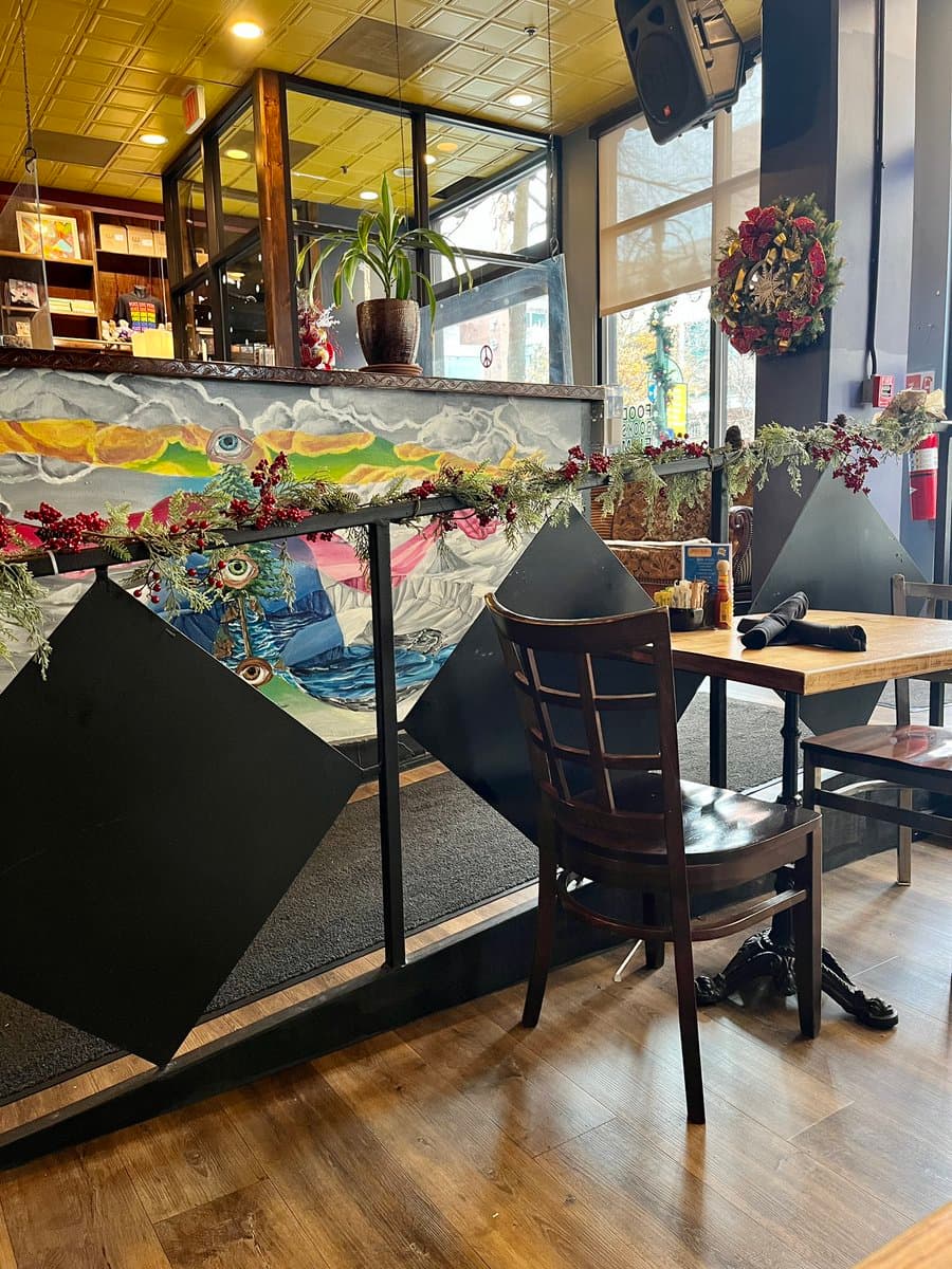 A photograph of a restaurant interior featuring a mural with multiple painted eyes on the wall, decorated with a garland of red berries and greenery. A wooden chair sits at a table in the foreground, and a Christmas wreath hangs on the window to the right.