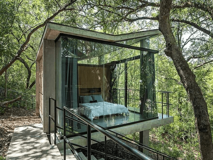 A modern glass-walled treehouse bedroom is nestled among tall trees, featuring a bed with light blue linens and a concrete platform with black railings. The structure is surrounded by dense green foliage and dappled sunlight filtering through the canopy.