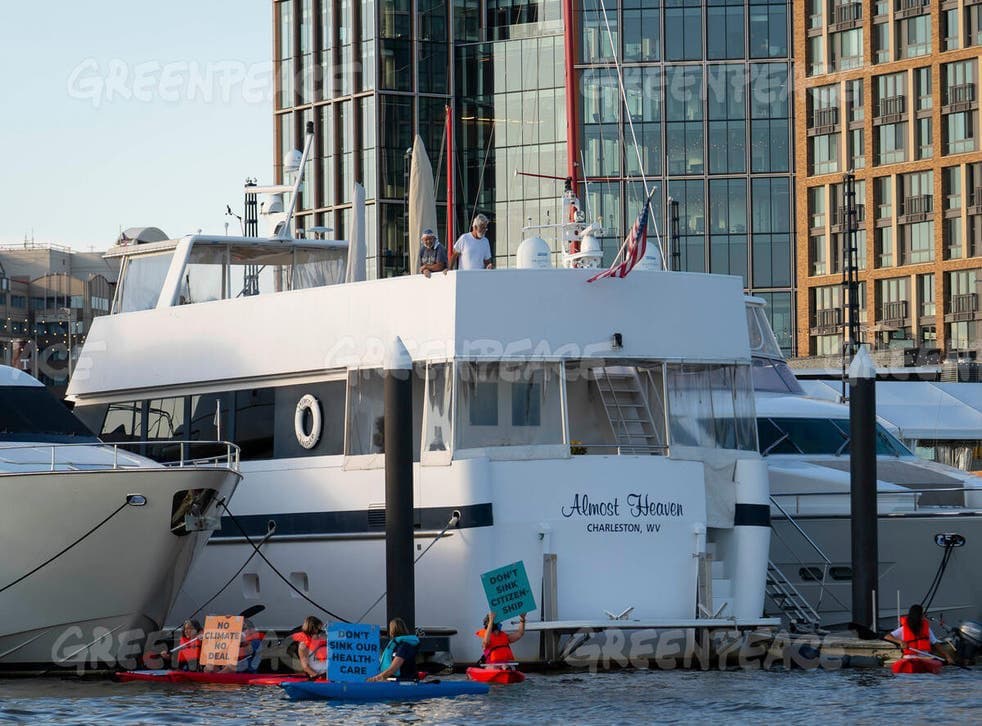 A large white yacht named 'Almost Heaven' is docked in a marina, with several people in kayaks holding protest signs in the foreground. The scene is set against a backdrop of modern high-rise buildings.