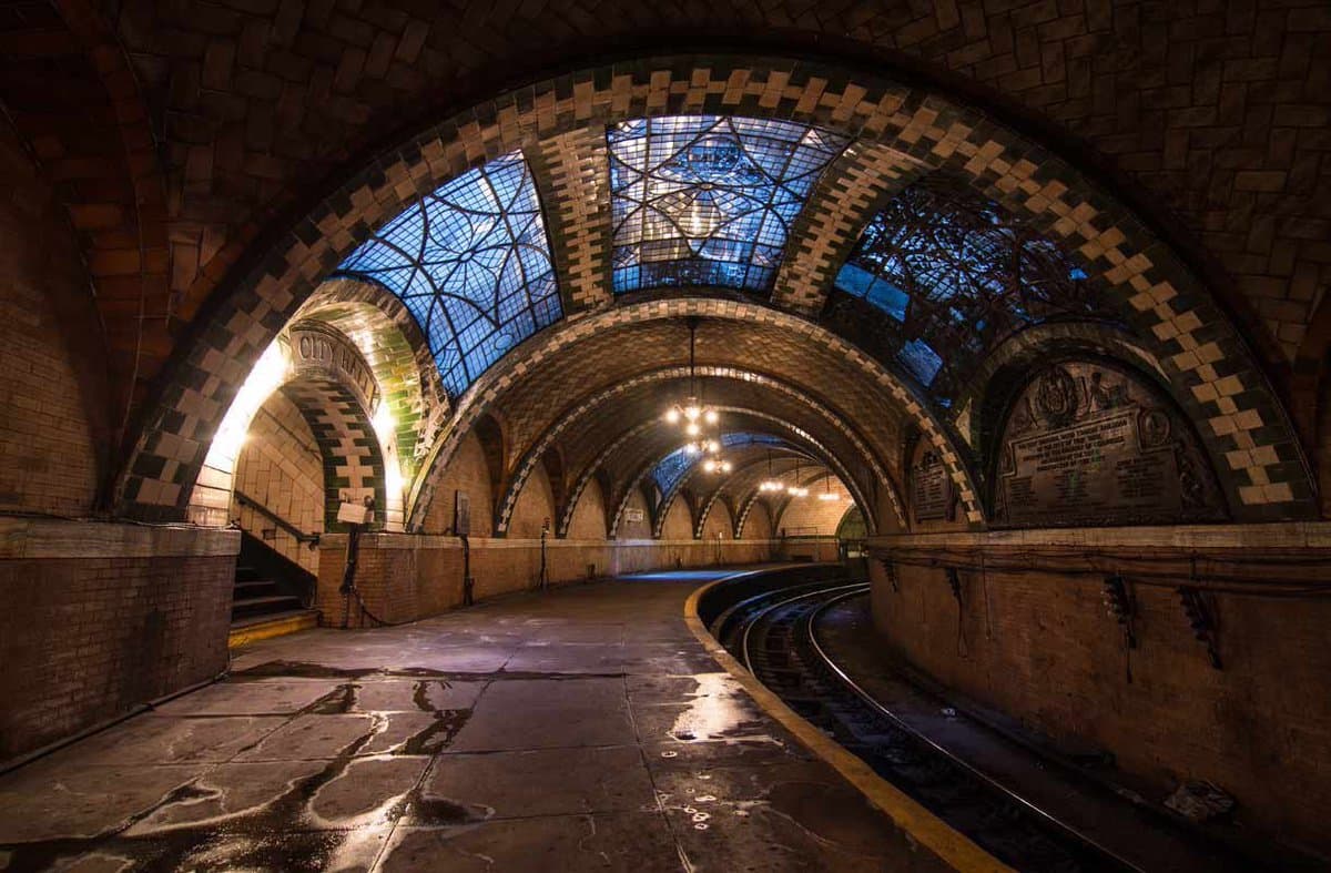 A wide-angle photograph of an abandoned, ornate subway station featuring a vaulted ceiling with stained-glass windows, tiled arches, and a curved track bed.