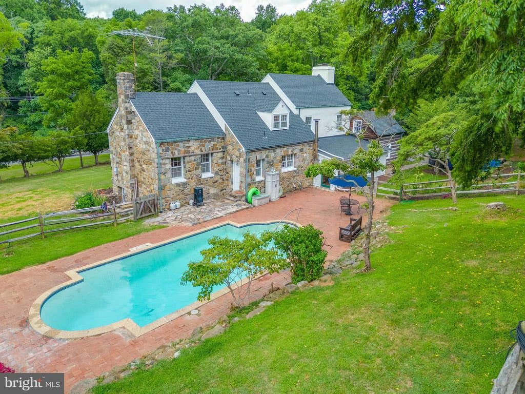 A real estate photograph showing a large, historic-style stone and white house with a dark roof, set on a grassy property with a swimming pool in the foreground. The image is a professional listing photo, likely taken from an elevated angle, featuring a 'bright MLS' watermark in the bottom left corner.