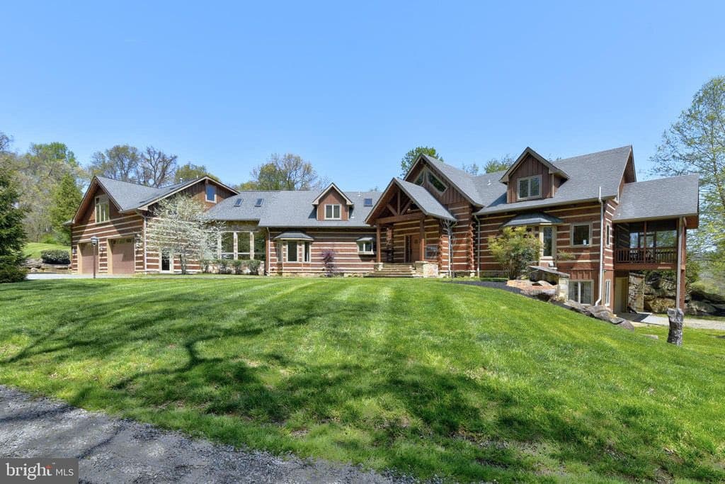 A wide exterior shot of a large, multi-story log cabin house with a gray shingled roof, set behind a large, manicured green lawn under a clear blue sky. The home features multiple gables, a covered porch, and a garage on the left side.
