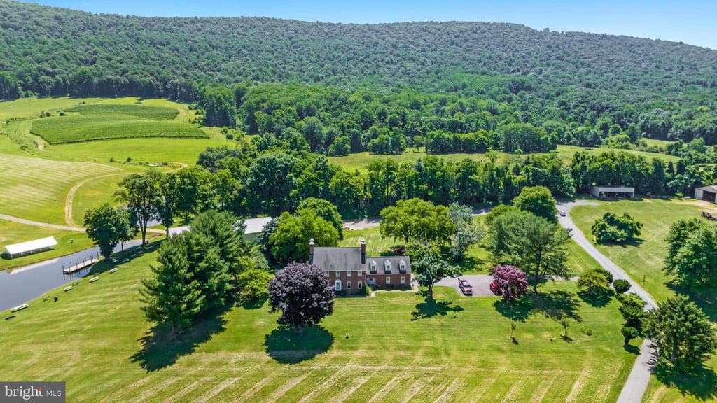 An aerial photograph of a large, historic-style brick house situated on a sprawling green estate with vineyards, a pond, and a forested hill in the background. The image is a real estate listing photo, as indicated by the watermark in the corner.