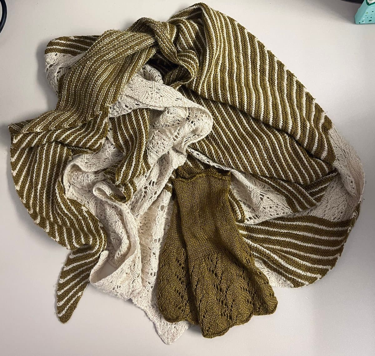 A top-down photograph of a hand-knitted shawl and a pair of fingerless mitts resting on a white surface. The shawl features a striped pattern in olive green and white, while the mitts are a solid olive green with a lace-like texture.