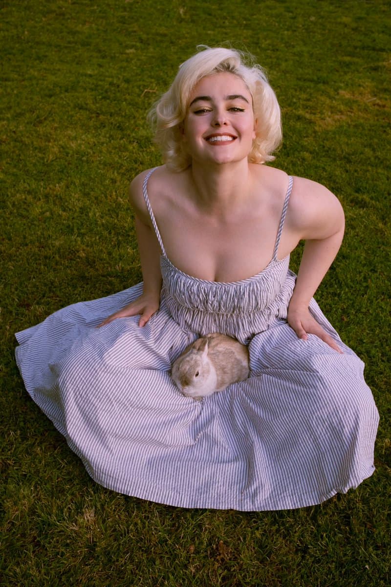 A woman with short blonde hair sits on a green lawn wearing a long, striped dress, smiling at the camera. A small white and brown rabbit is sitting on her lap.