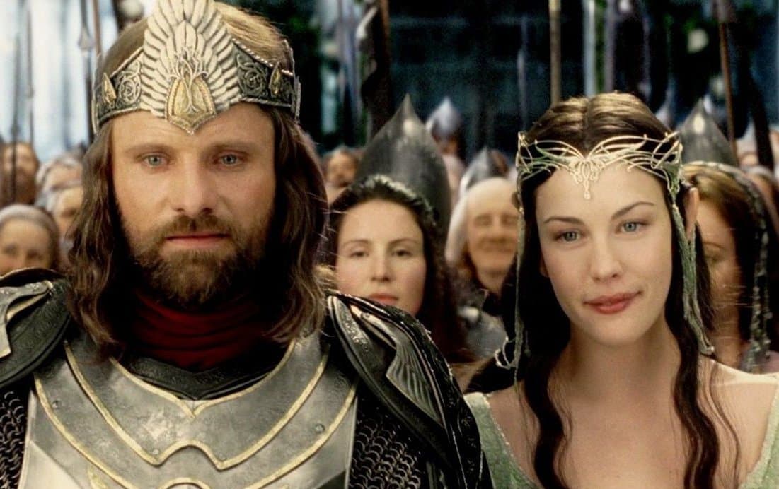 A still from a fantasy film featuring a bearded man in ornate silver armor and a crown standing next to a woman wearing a delicate silver headpiece, with a crowd of people in the background.
