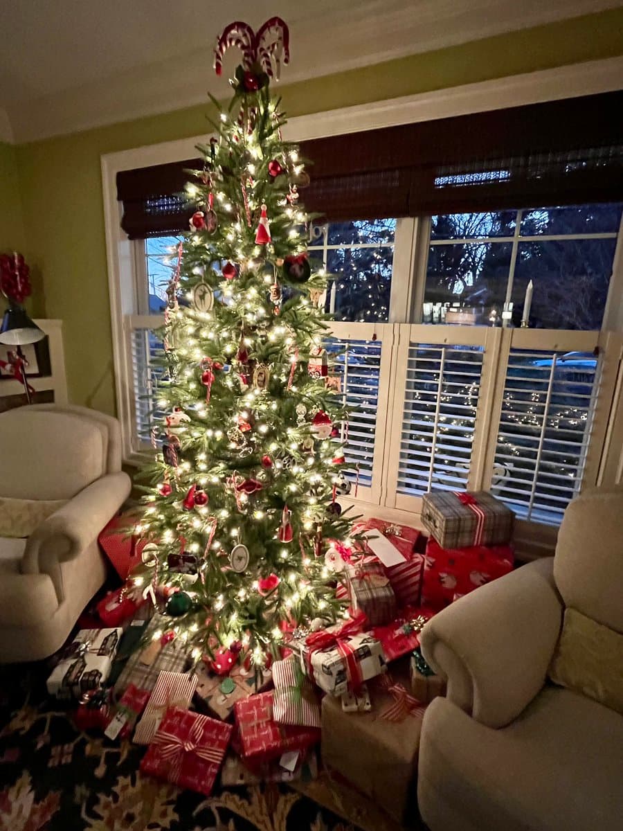 A decorated Christmas tree stands in a living room, illuminated by white string lights and topped with a candy cane topper. Numerous wrapped presents are piled on the floor around the base of the tree, and the room is furnished with beige armchairs and a patterned rug.