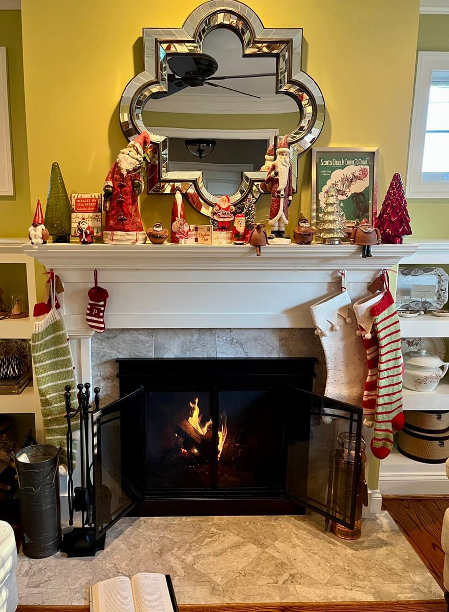A festive indoor scene featuring a lit fireplace with a marble hearth, decorated with Christmas stockings and a collection of Santa figurines and ornaments on the mantel. A large, ornate mirror hangs on the yellow wall above the mantel, reflecting a ceiling fan.