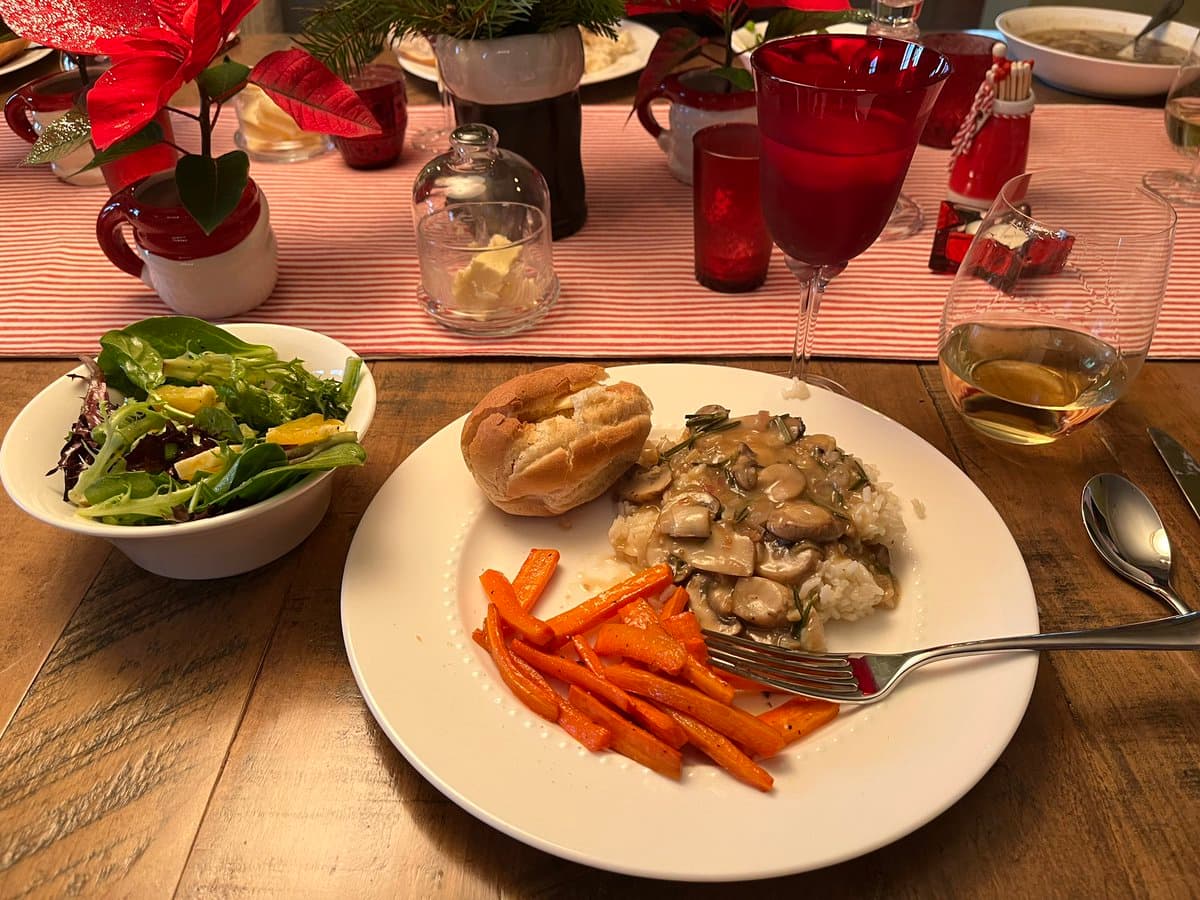 A close-up photograph of a festive dinner setting featuring a plate with a mushroom and rice dish, roasted carrots, and a bread roll, accompanied by a side salad and red holiday decorations.