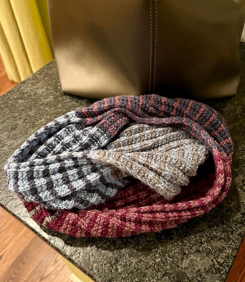 A photograph of three hand-knitted scarves in different colorways, coiled together on a speckled countertop. The scarves feature textured stitches in shades of gray, white, beige, and maroon.