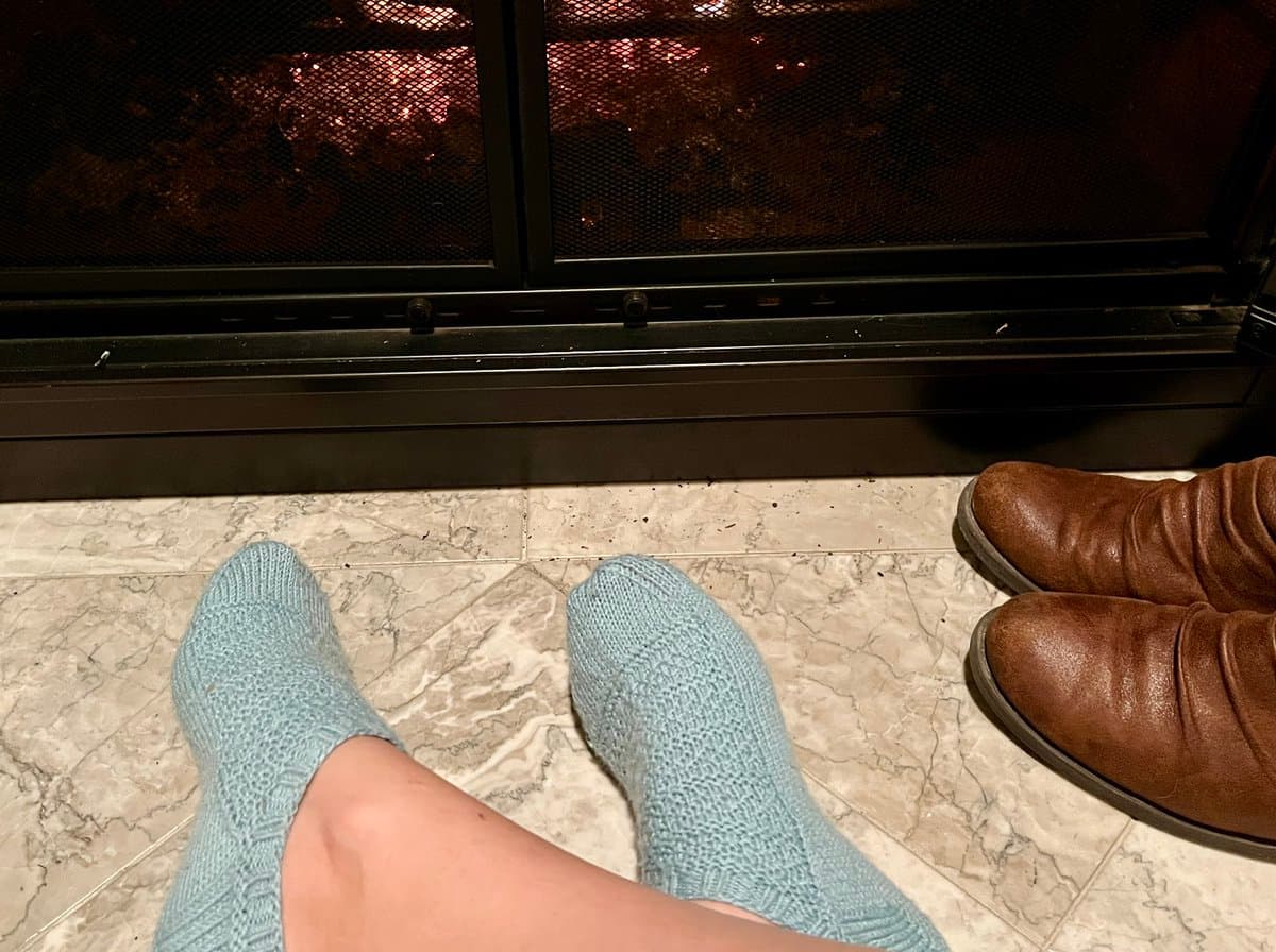 A first-person view of a person's feet wearing light blue hand-knitted socks, resting on a tiled floor in front of a fireplace. A pair of brown leather boots sits nearby on the right.