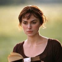 A medium shot of a woman with brown hair tied back, wearing a dark brown top, holding an open book in her hands against a blurred green background.