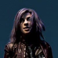 A person with long, dark hair and a leather jacket is shown in a dimly lit setting, looking slightly to the side with a serious expression. The image appears to be a photograph of a musician or performer on stage.